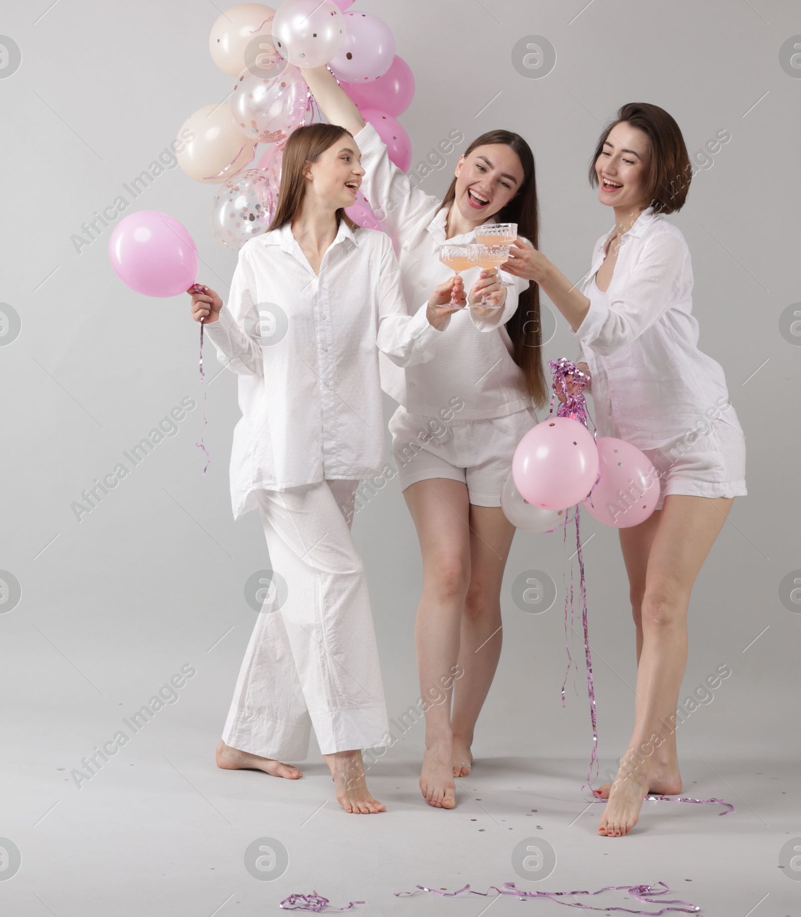 Photo of Pajama party. Happy friends with tinsel and balloons clinking glasses on grey background