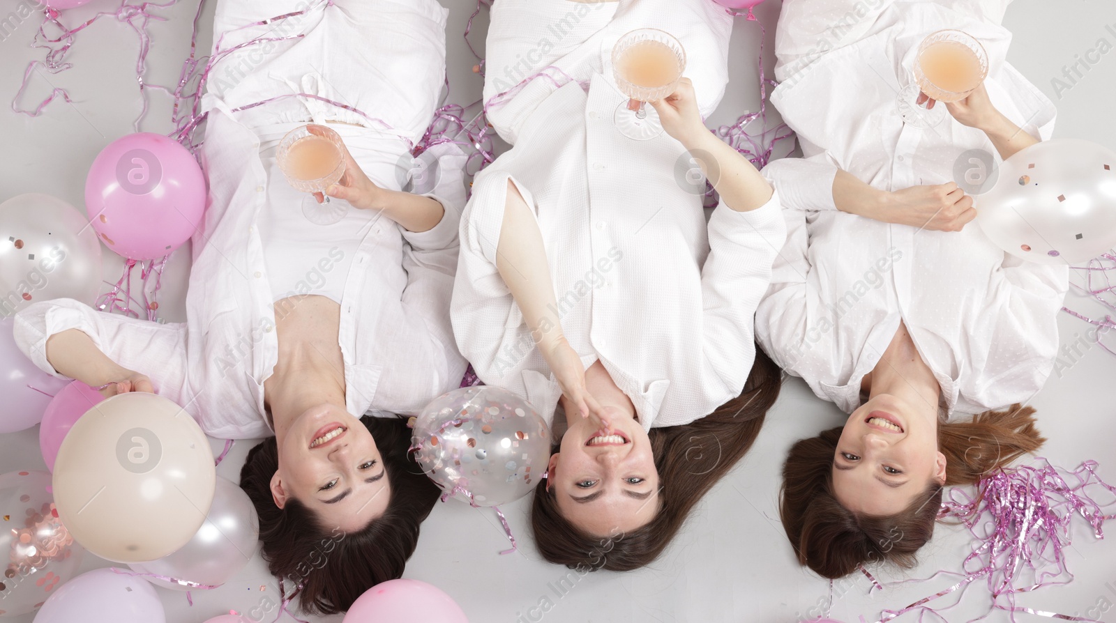 Photo of Pajama party. Happy friends with glasses of alcoholic drink, balloons and tinsel on grey background, top view