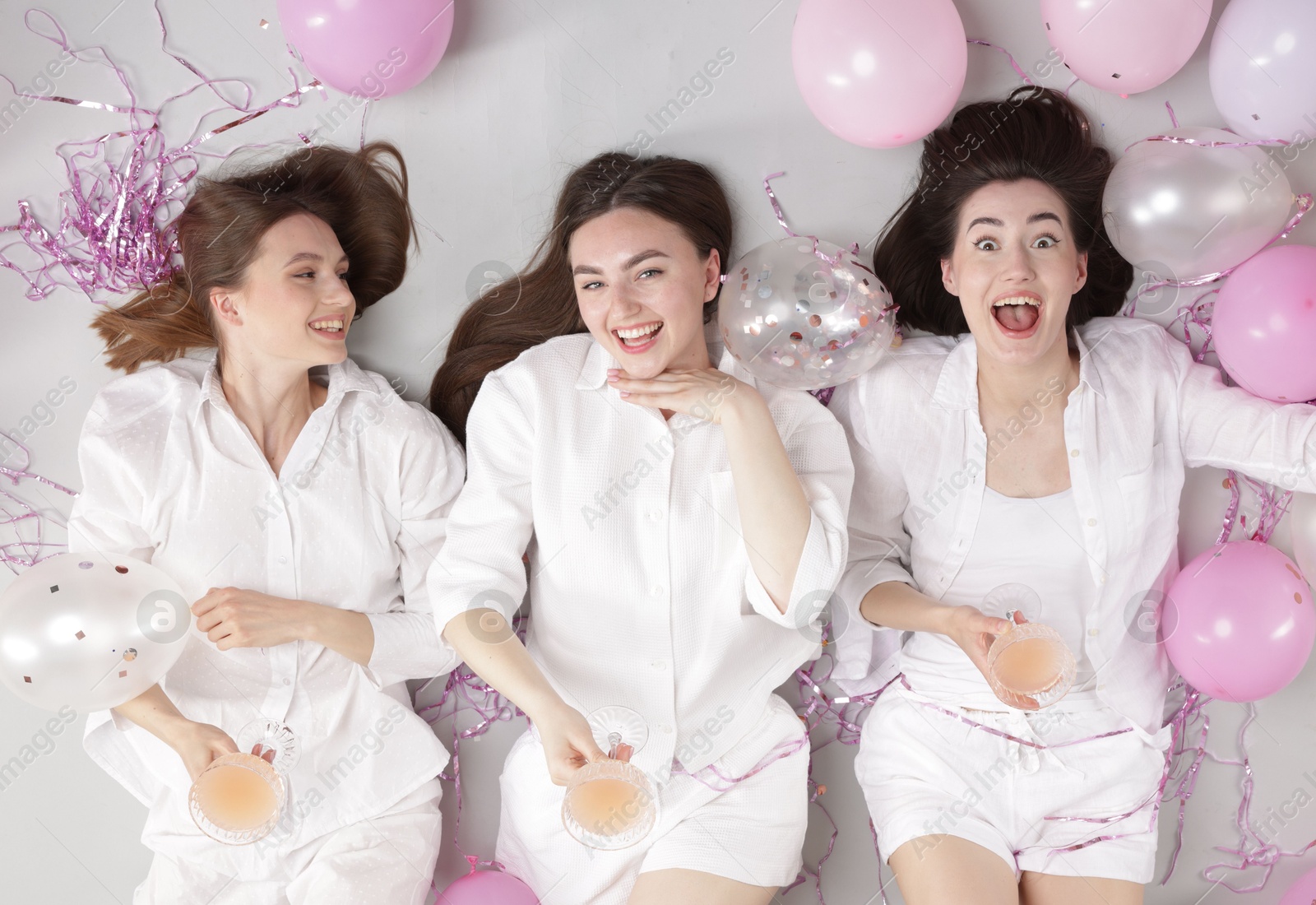 Photo of Pajama party. Happy friends with glasses of alcoholic drink, balloons and tinsel on grey background, top view