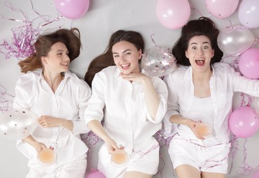 Photo of Pajama party. Happy friends with glasses of alcoholic drink, balloons and tinsel on grey background, top view