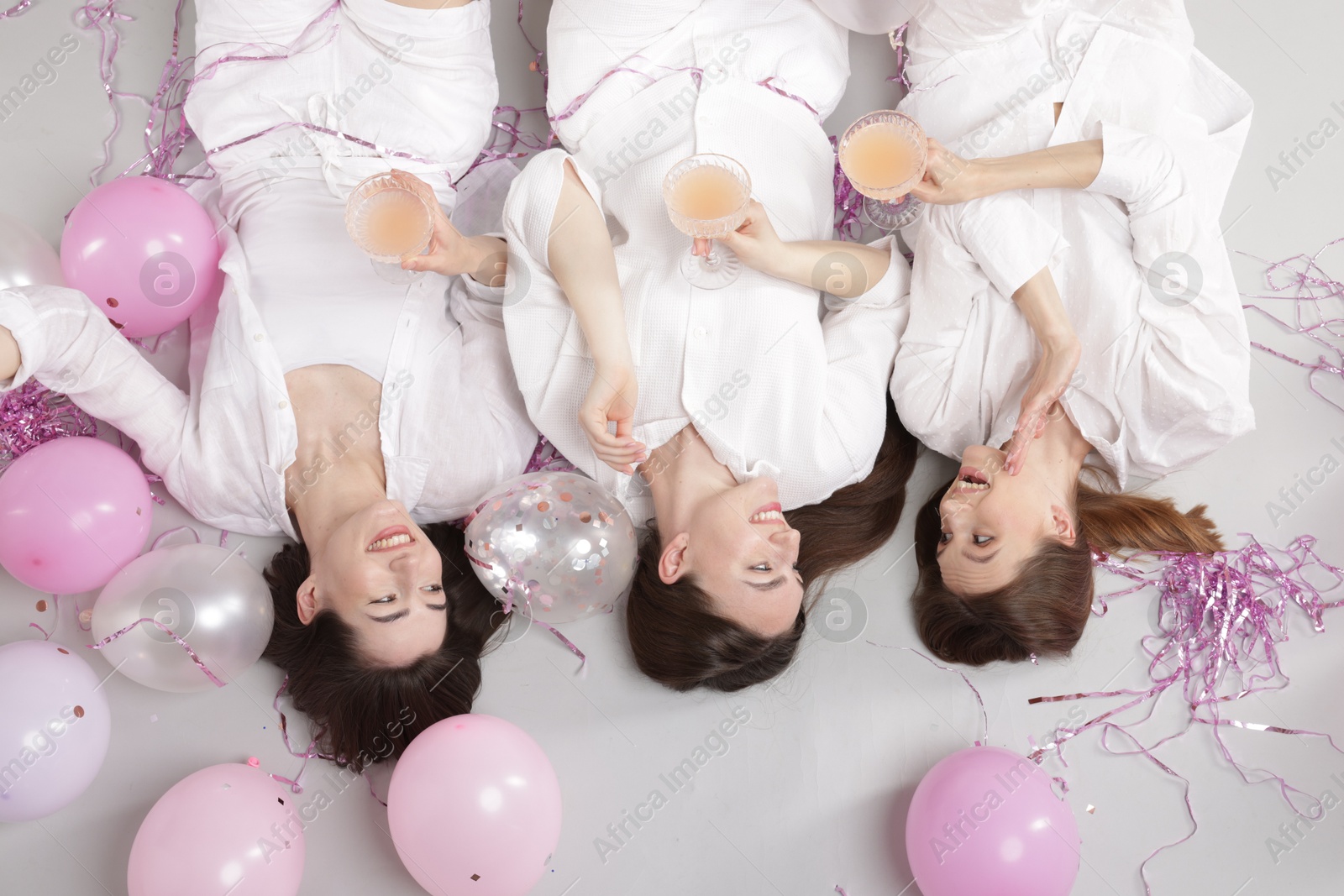 Photo of Pajama party. Happy friends with glasses of alcoholic drink, balloons and tinsel on grey background, top view