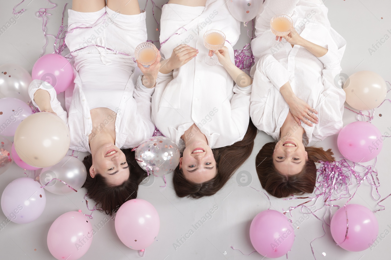 Photo of Pajama party. Happy friends with glasses of alcoholic drink, balloons and tinsel on grey background, top view