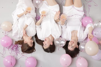 Photo of Pajama party. Happy friends with glasses of alcoholic drink, balloons and tinsel on grey background, top view