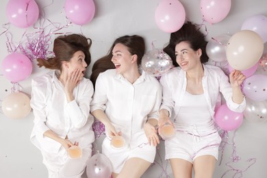 Photo of Pajama party. Happy friends with glasses of alcoholic drink, balloons and tinsel on grey background, top view