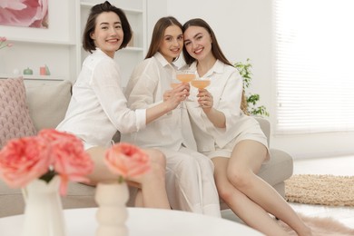 Photo of Pajama party. Happy friends clinking glasses on sofa at home