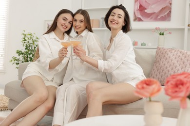 Photo of Pajama party. Happy friends clinking glasses on sofa at home