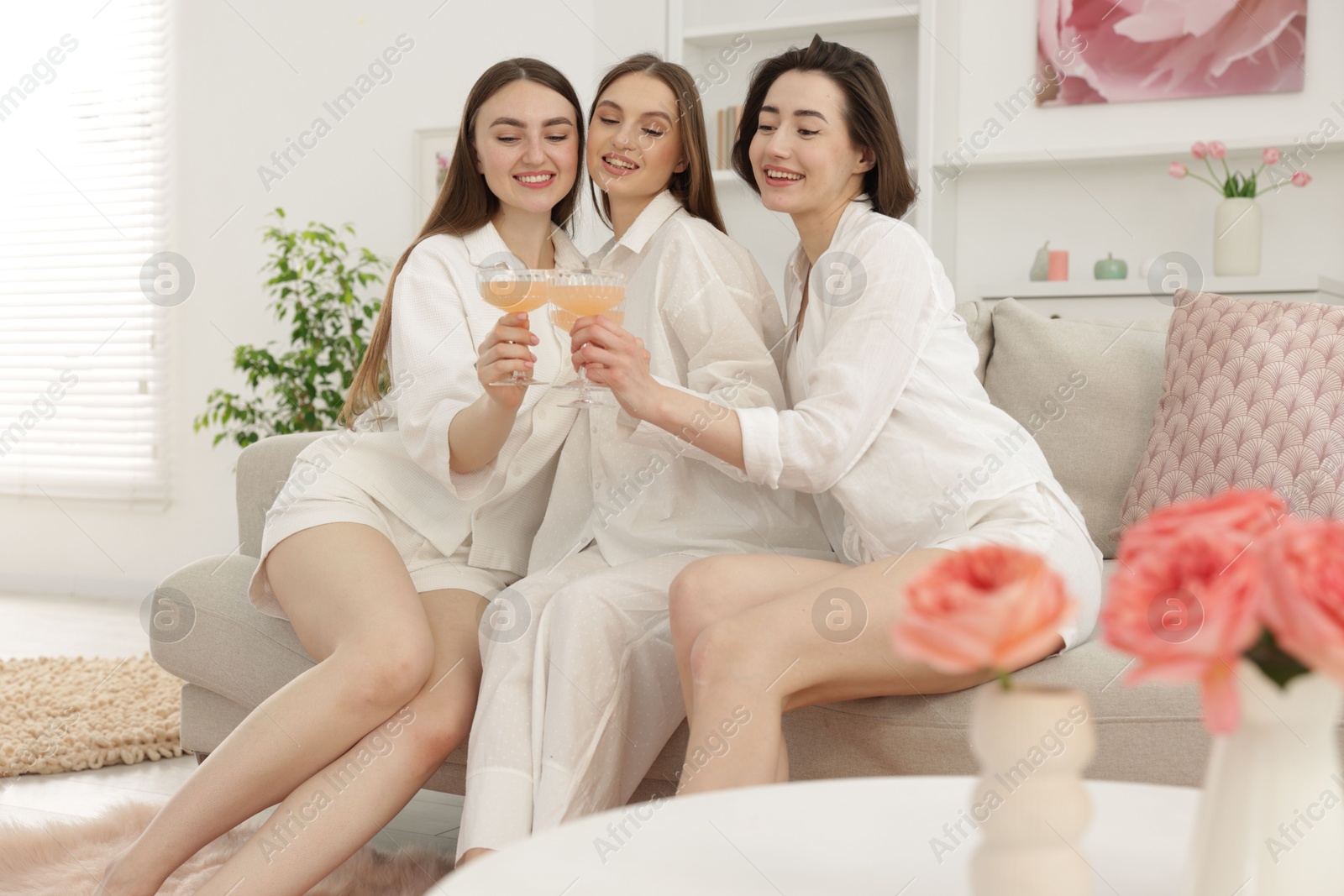 Photo of Pajama party. Happy friends clinking glasses on sofa at home