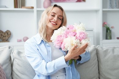Photo of Happy woman with bouquet of beautiful peonies on sofa at home