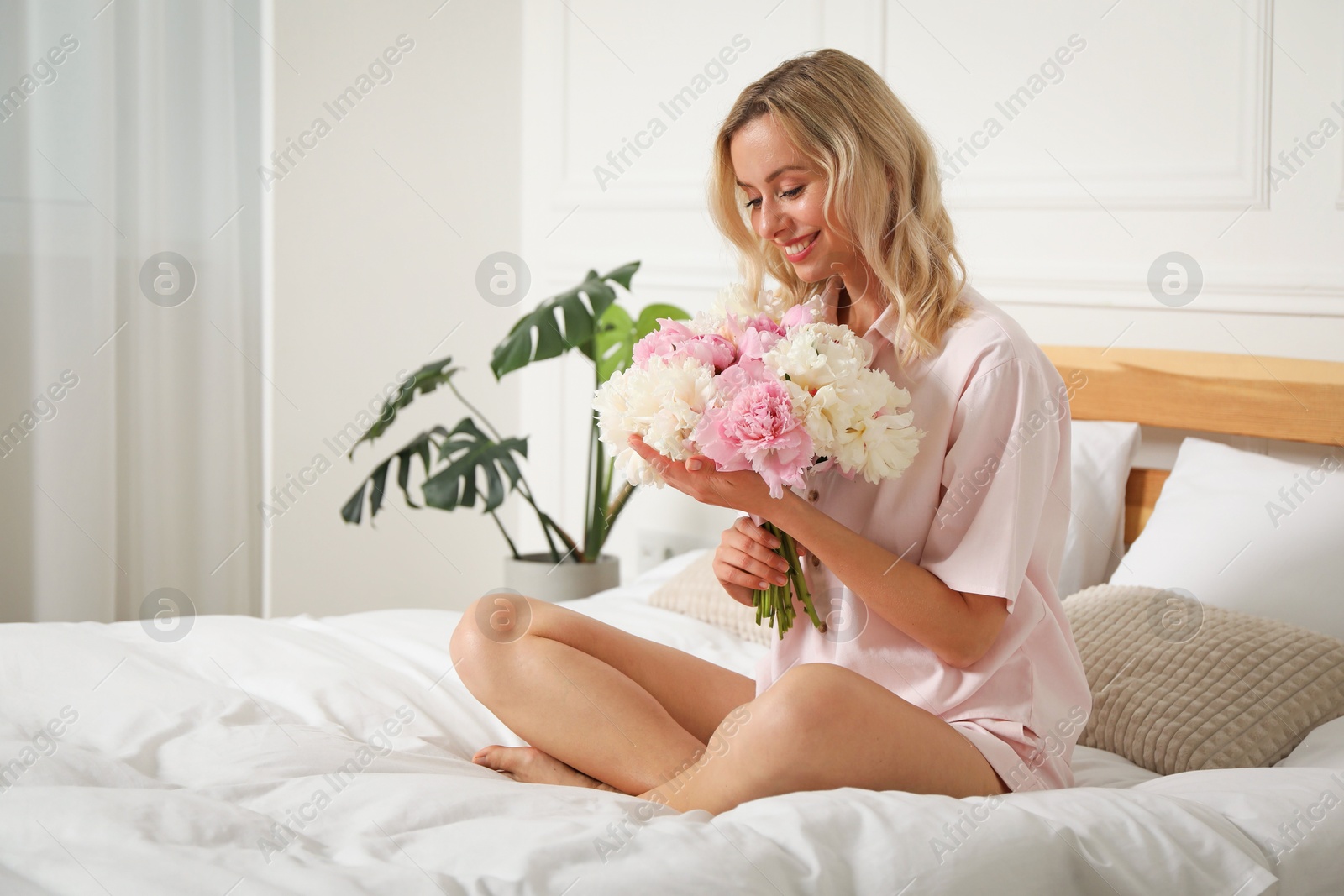 Photo of Happy woman with bouquet of beautiful peonies on bed indoors, space for text