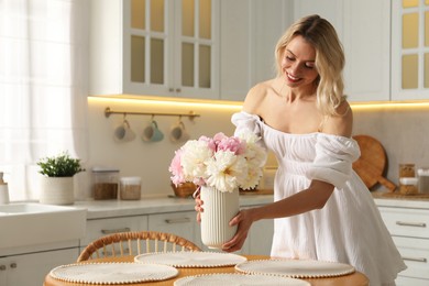 Photo of Happy woman arranging vase with bouquet of beautiful peonies onto table in kitchen