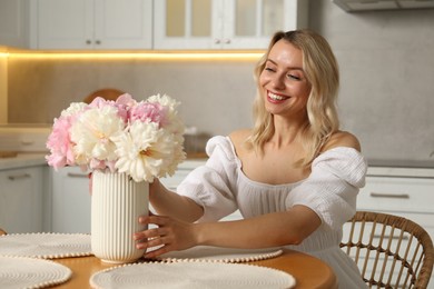 Photo of Happy woman with bouquet of beautiful peonies at table in kitchen