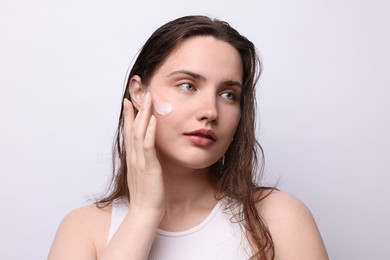 Photo of Beautiful woman applying skincare product onto her face on light grey background, closeup