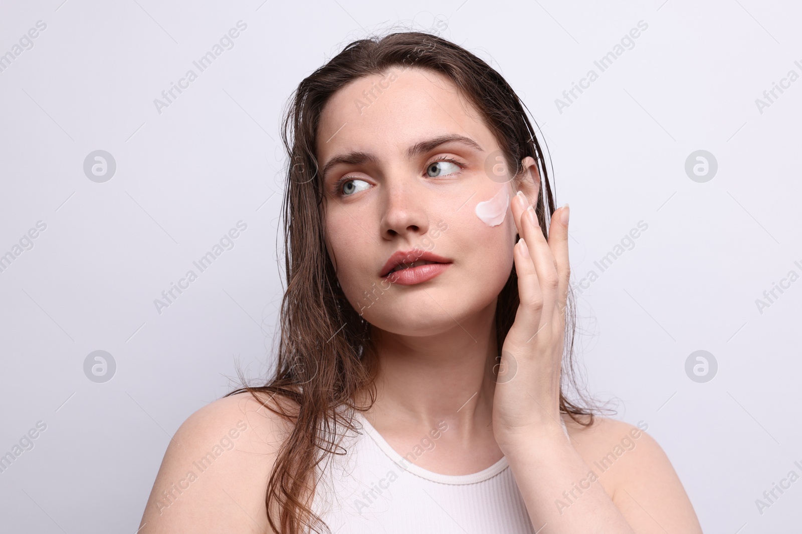 Photo of Beautiful woman applying skincare product onto her face on light grey background, closeup