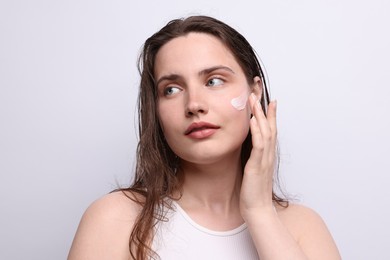 Photo of Beautiful woman applying skincare product onto her face on light grey background, closeup