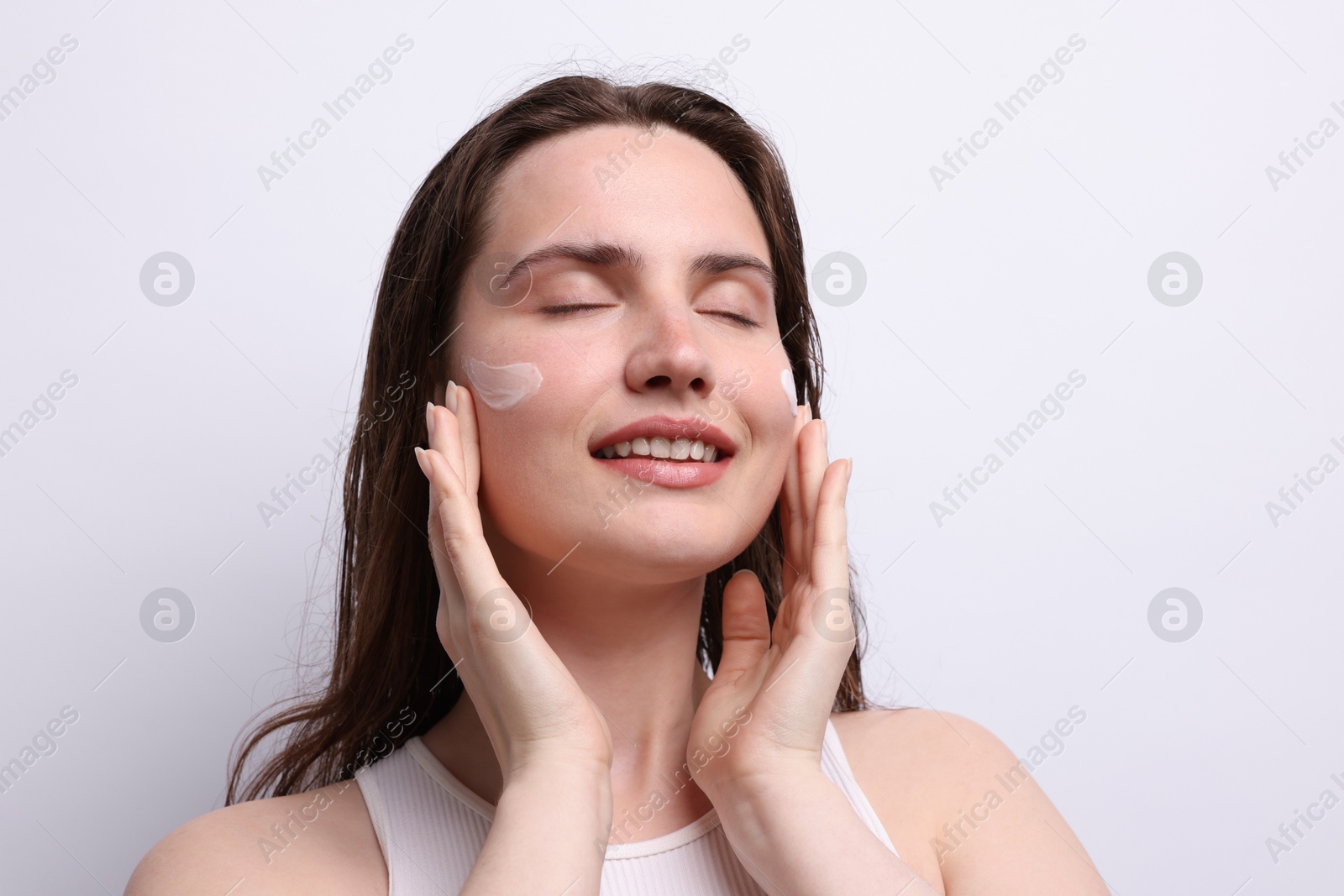 Photo of Smiling woman applying skincare product onto her face on light grey background, closeup