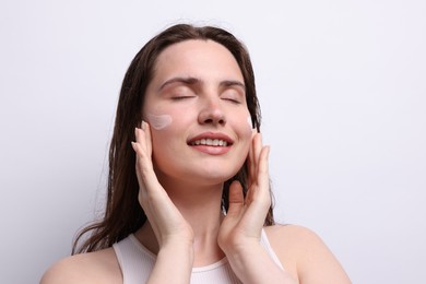 Photo of Smiling woman applying skincare product onto her face on light grey background, closeup