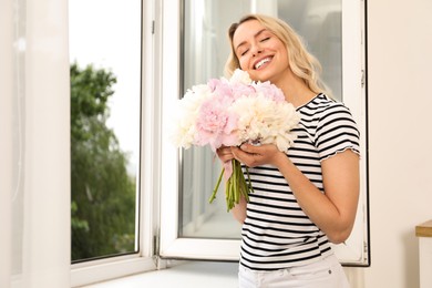 Photo of Happy woman with bouquet of beautiful peonies near window indoors, space for text