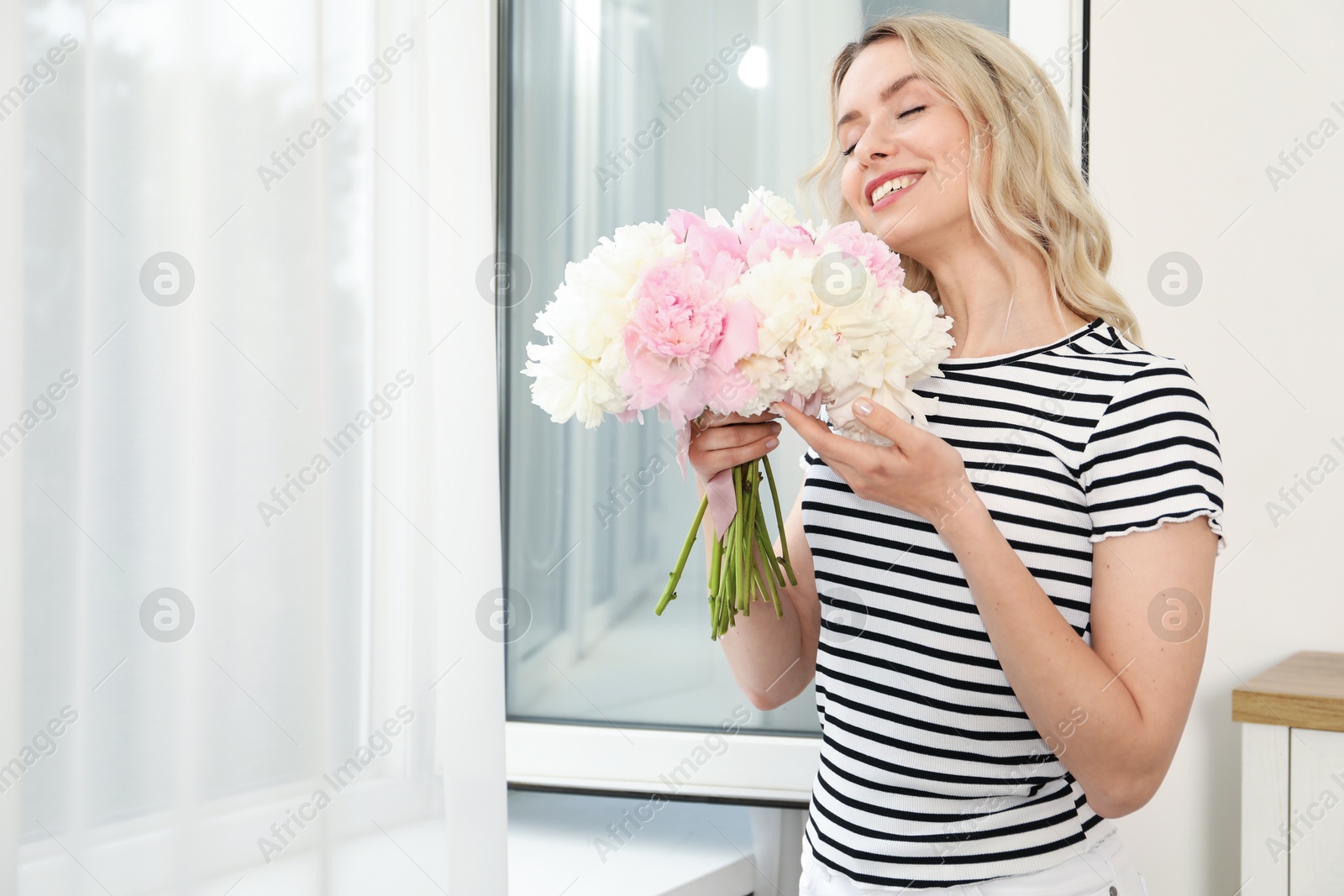 Photo of Happy woman with bouquet of beautiful peonies near window indoors