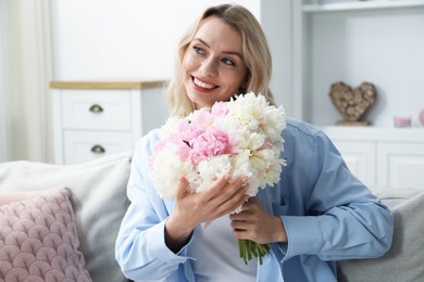 Photo of Happy woman with bouquet of beautiful peonies on sofa at home