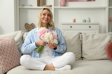 Photo of Happy woman with bouquet of beautiful peonies on sofa at home