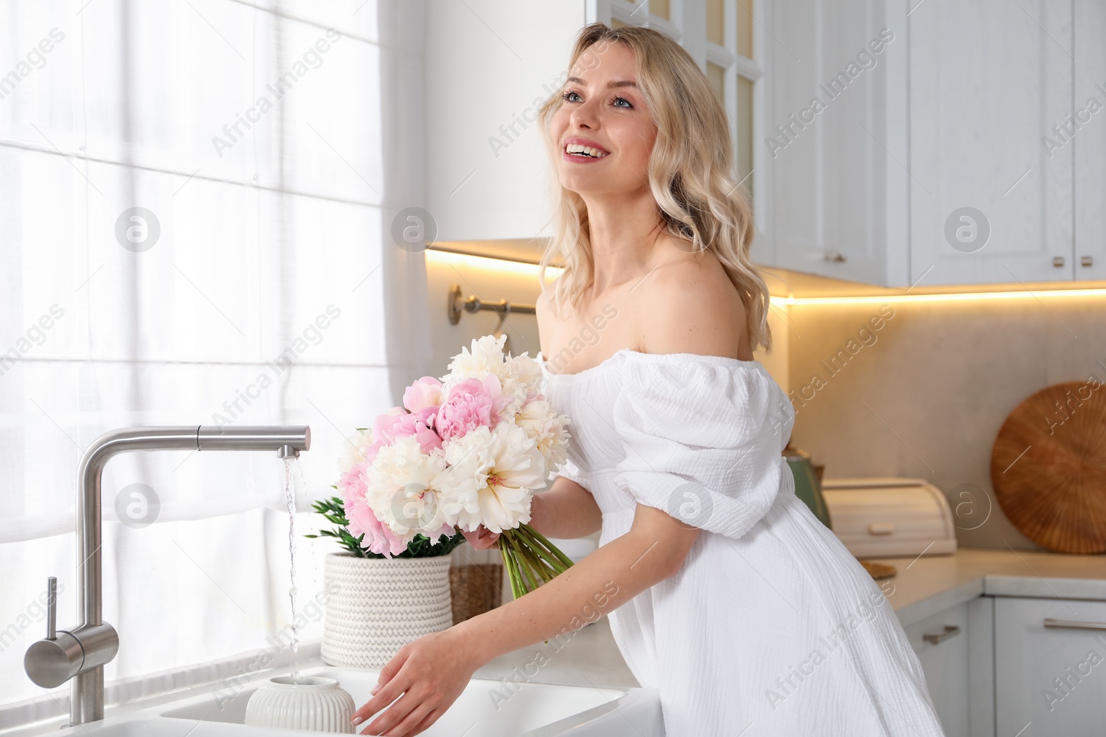 Photo of Happy woman taking care of beautiful peonies in kitchen