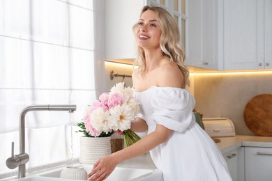 Photo of Happy woman taking care of beautiful peonies in kitchen