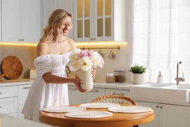 Photo of Happy woman arranging vase with bouquet of beautiful peonies onto table in kitchen