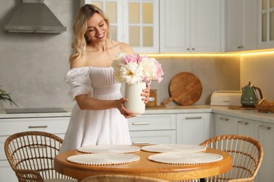 Photo of Happy woman arranging vase with bouquet of beautiful peonies onto table in kitchen