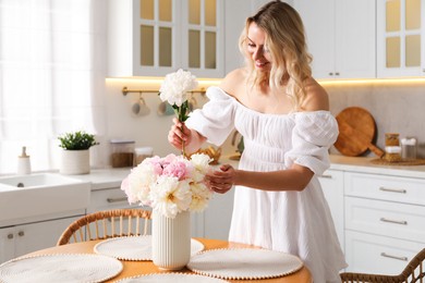 Photo of Happy woman making bouquet of beautiful peonies at table in kitchen