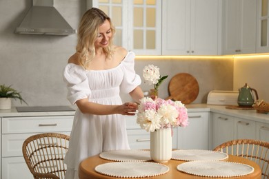 Photo of Happy woman making bouquet of beautiful peonies at table in kitchen