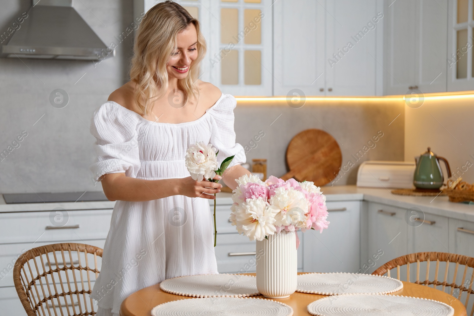 Photo of Happy woman making bouquet of beautiful peonies at table in kitchen