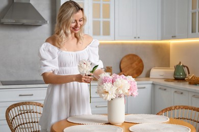 Photo of Happy woman making bouquet of beautiful peonies at table in kitchen
