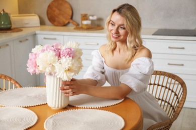 Photo of Happy woman with bouquet of beautiful peonies at table in kitchen