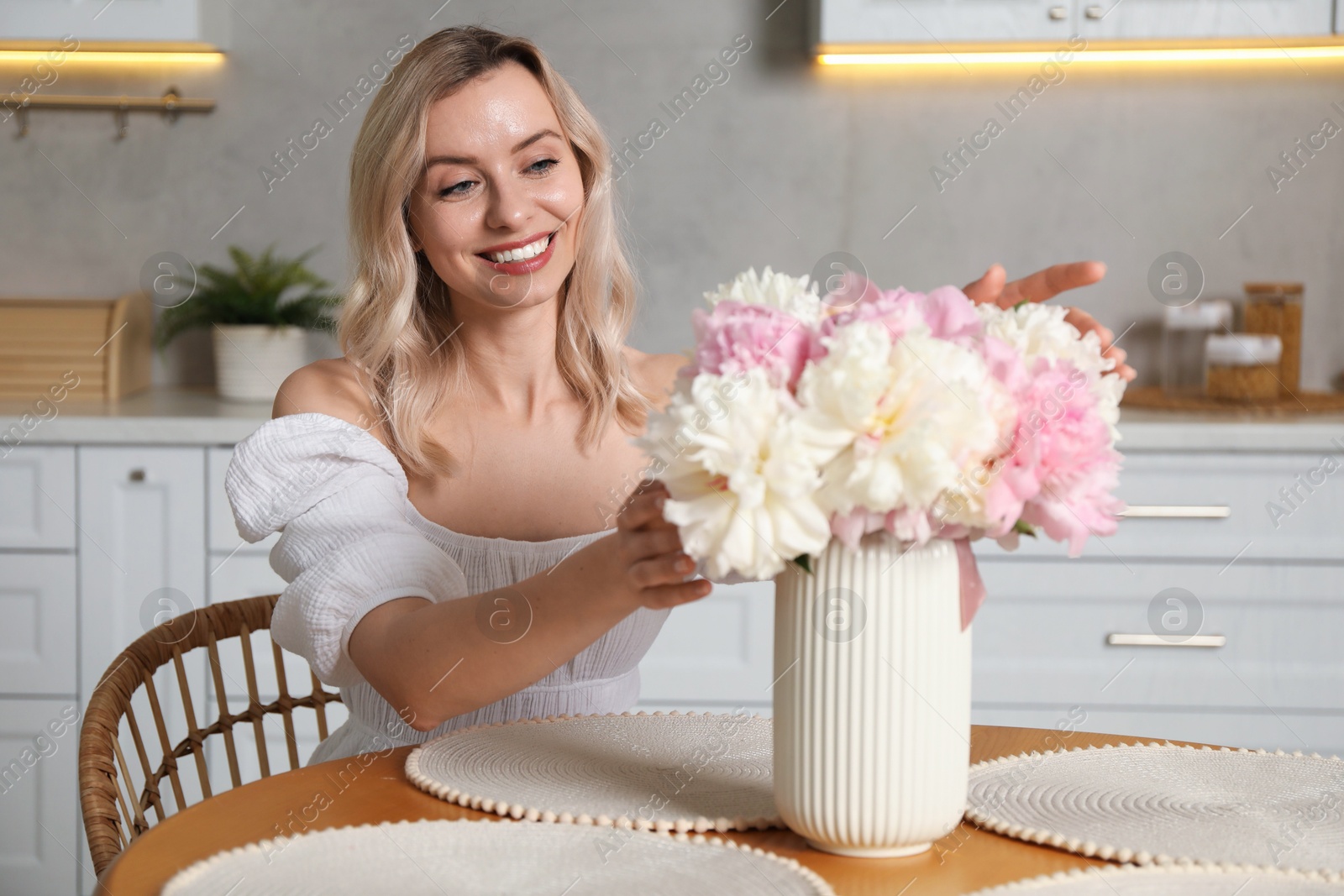 Photo of Happy woman with bouquet of beautiful peonies at table in kitchen