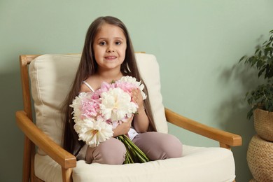 Photo of Cute little girl with bouquet of beautiful peonies in armchair near green wall indoors