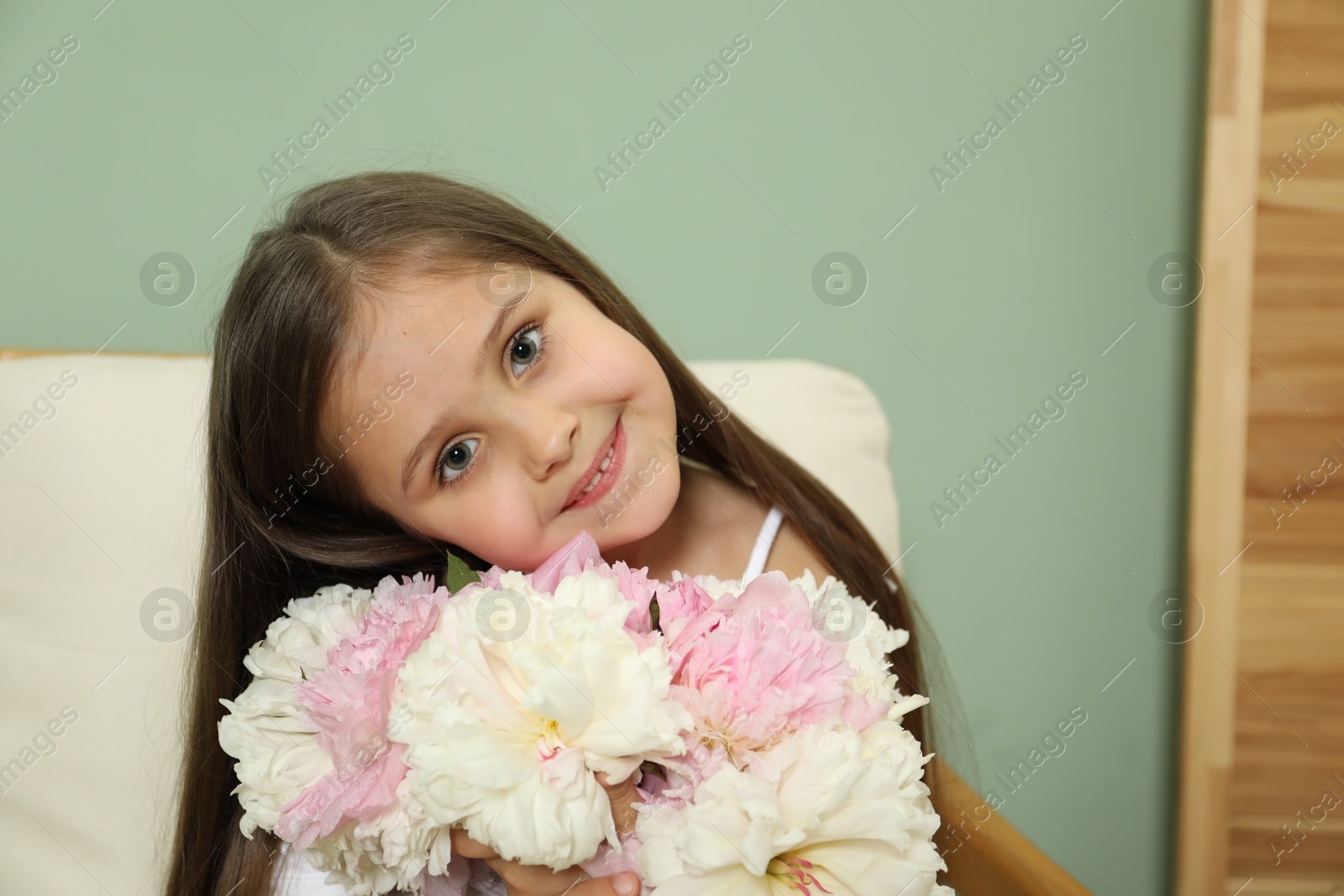 Photo of Cute little girl with bouquet of beautiful peonies in armchair near green wall indoors