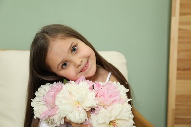 Photo of Cute little girl with bouquet of beautiful peonies in armchair near green wall indoors