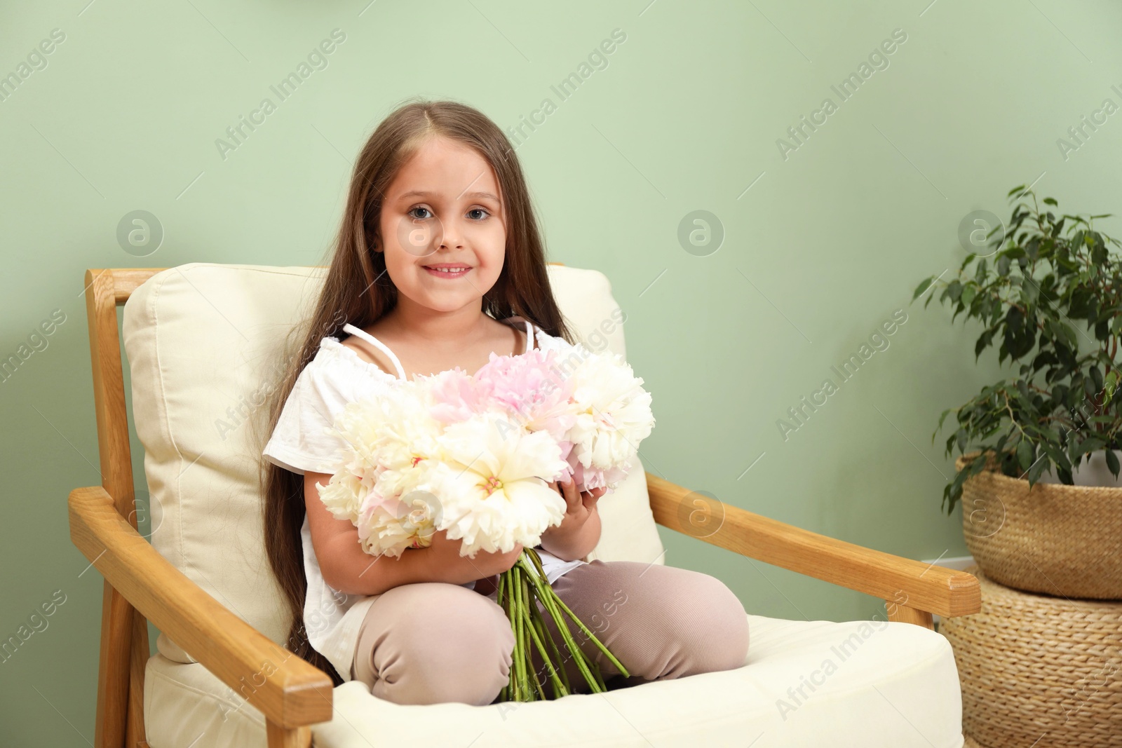 Photo of Cute little girl with bouquet of beautiful peonies in armchair near green wall indoors