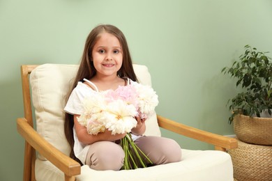 Photo of Cute little girl with bouquet of beautiful peonies in armchair near green wall indoors