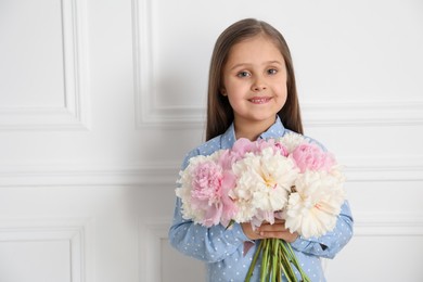 Photo of Cute little girl with bouquet of beautiful peonies near white wall indoors, space for text