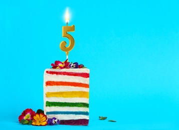 Image of Piece of delicious rainbow cake with burning golden candle in shape of number 5 and flowers on light blue background, space for text. Birthday or other special occasion