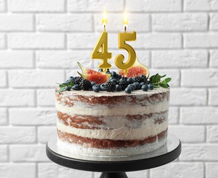 Image of Yummy cake with fruits and burning number shaped candles near white brick wall. 45th birthday or other special occasion