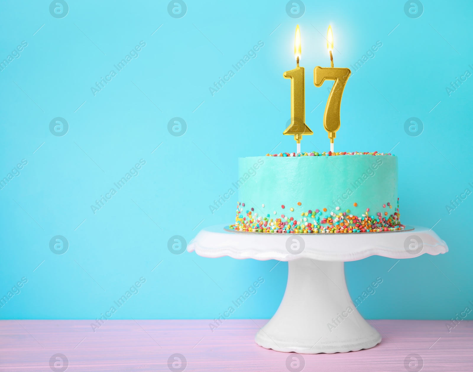 Image of Yummy cake with burning number shaped candles and sprinkles on white table against turquoise background, space for text. 17th birthday or other special occasion