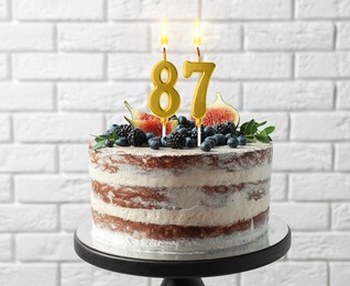 Image of Yummy cake with fruits and burning number shaped candles near white brick wall. 87th birthday or other special occasion