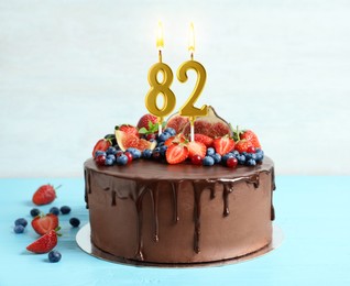 Image of Yummy chocolate cake with fruits and burning number shaped candles on light blue table. 82th birthday or other special occasion