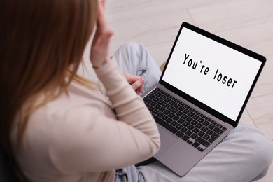 Photo of Cyber bullying. Upset teenage girl looking at laptop with text You Loser on floor indoors, closeup