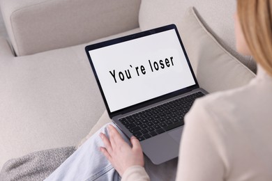 Photo of Cyber bullying. Upset teenage girl looking at laptop with text You Loser on sofa at home, closeup