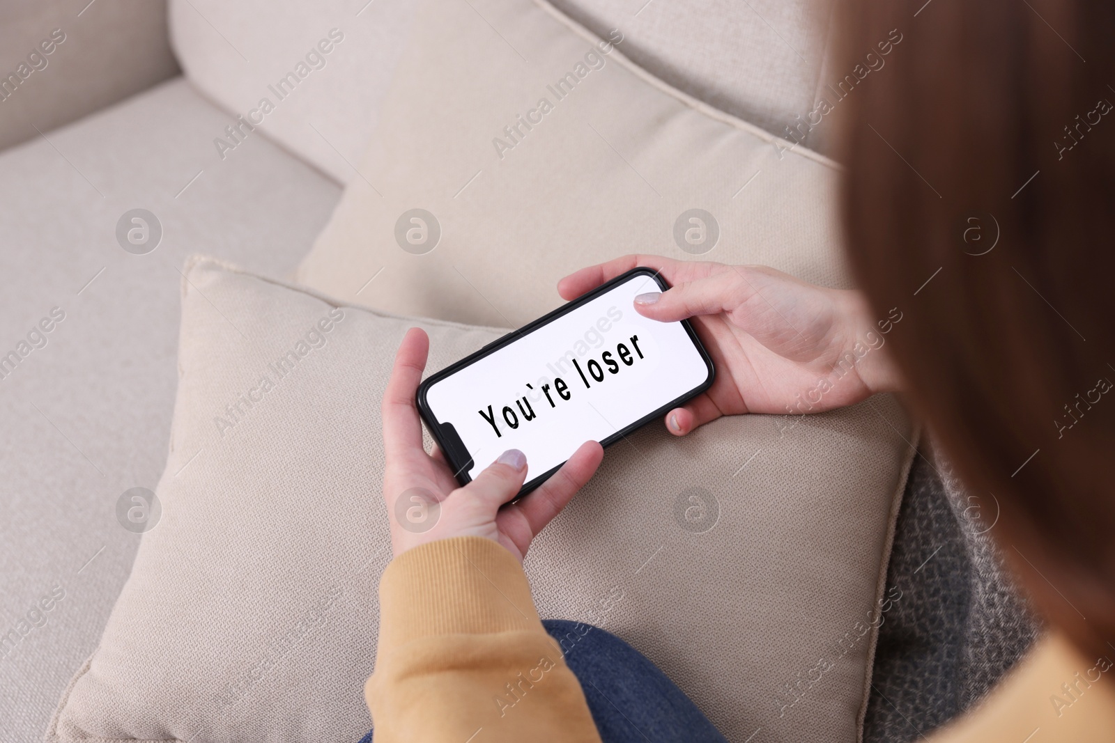 Cyber bullying. Upset teenage girl looking at smartphone with text You Loser on sofa at home, closeup Photo of Cyber bullying. Upset teenage girl looking at smartphone with text You Loser on sofa at home, closeup