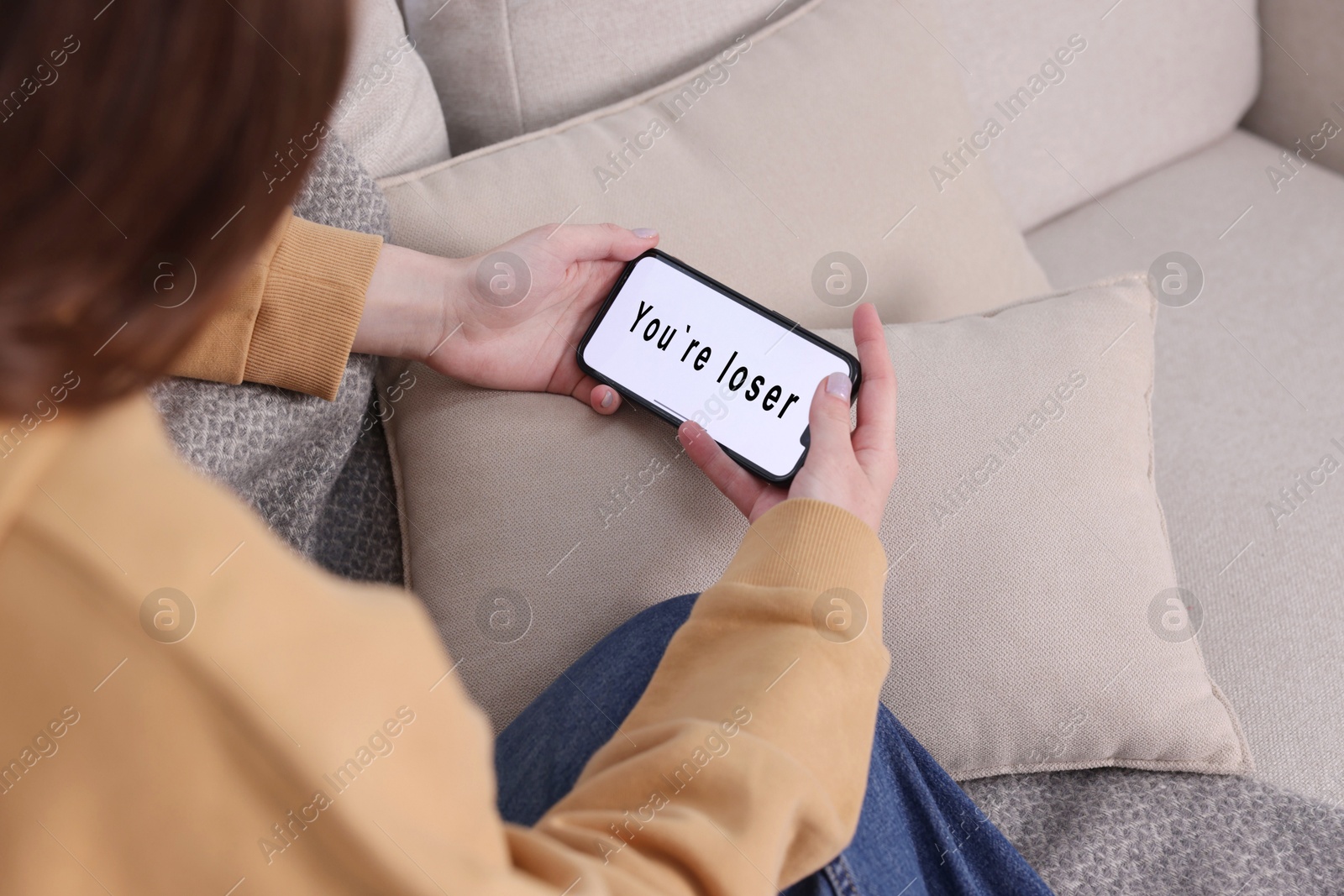 Cyber bullying. Upset teenage girl looking at smartphone with text You Loser on sofa at home, closeup Photo of Cyber bullying. Upset teenage girl looking at smartphone with text You Loser on sofa at home, closeup