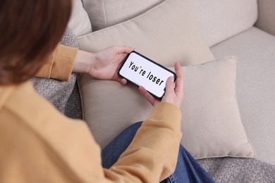 Photo of Cyber bullying. Upset teenage girl looking at smartphone with text You Loser on sofa at home, closeup